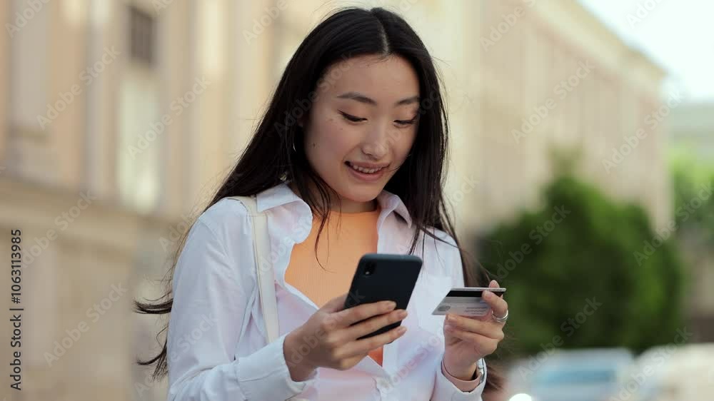 Asian woman makes online payments through the Internet from bank card on smartphone. Student girl on street with credit card and phone. Woman celebrating a successful purchase. Asian woman makes online payments through the Internet from bank card on smartphone. Student girl on street with credit card and phone. Woman celebrating a successful purchase.