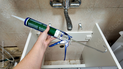 A person's hand holds a caulking gun over a bathroom sink cabinet, illustrating home repair and plumbing maintenance