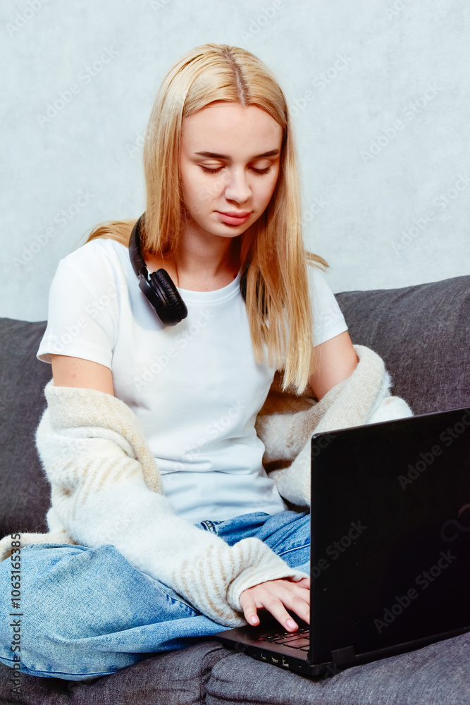 Naklejka premium A young woman works in the living room, sitting on the sofa with headphones and a laptop. A cozy moment that highlights the comfort of remote work and focus on tasks.