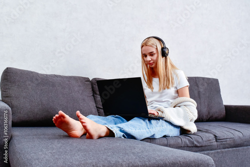 A young woman works in the living room, sitting on the sofa with headphones and a laptop. A cozy moment that highlights the comfort of remote work and focus on tasks.