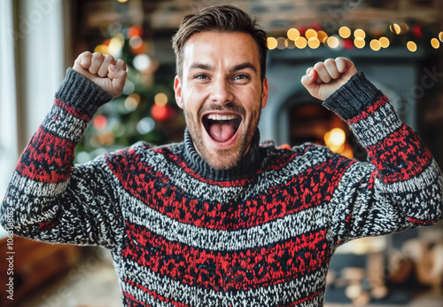 Happy man wearing christmas sweater raising fists and shouting with joy in front of christmas tree and fireplace photo