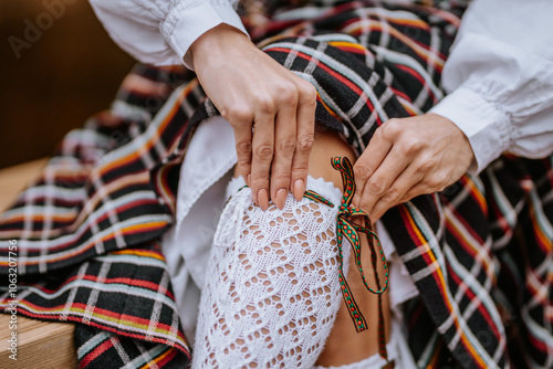 Close-up of hands adjusting a white lace stocking with a colorful ribbon over a traditional Latvian checkered skirt, showing detailed folk attire.. photo