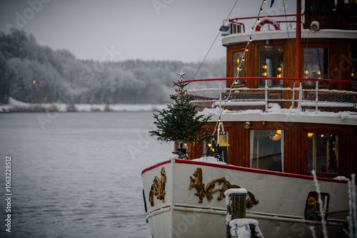 Decorated boat with a Christmas tree standing at edge of bow and warm lights, docked in snowy harbor, with its wooden cabin contrasting icy waters, creating cozy atmosphere for winter season.