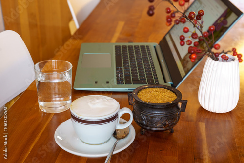 Break at work. Home office scene. A notebook, a glass of water, a cup of cappuccino, a sugar bowl and a small vase with a branch of the ornamental apple (Malus floribunda) on a wooden desk photo