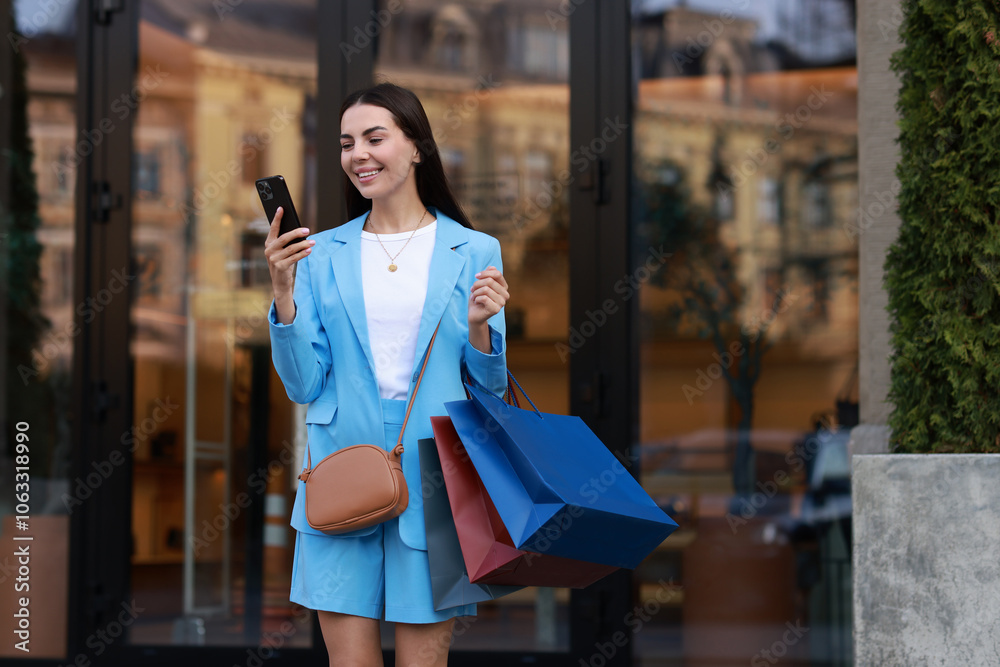Obraz premium Happy woman with colorful shopping bags using smartphone outdoors