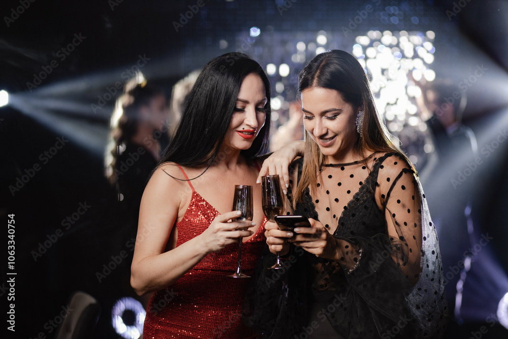 two women at a New Year or Christmas party, dressed in elegant dresses, holding glasses of champagne and smiling while looking at the phone screen. The atmosphere of the party is filled with warmth an