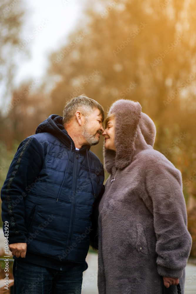 Fototapeta premium Elderly couple strolling hand in hand in a picturesque autumn park, sharing tender kisses amidst the colorful leaves, embodying enduring love and joy