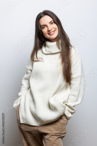 Portrait of a beautiful young woman wearing a white sweater and beige pants, on light gray background.