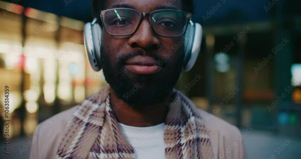 Camera moving closer to African American mans face. Male with glasses looking at camera and smiling. Putting on new headphones. Listening to music while spending free time in city.