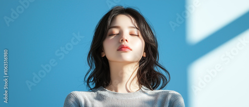 woman with closed eyes stands against blue background, basking in sunlight. Her serene expression and casual attire create peaceful atmosphere