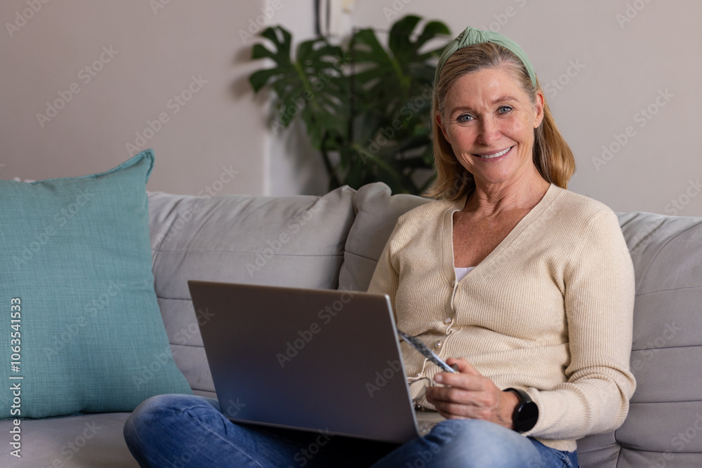 senior woman using laptop on couch at home, smiling and holding glasses senior woman using laptop on couch at home, smiling and holding glasses