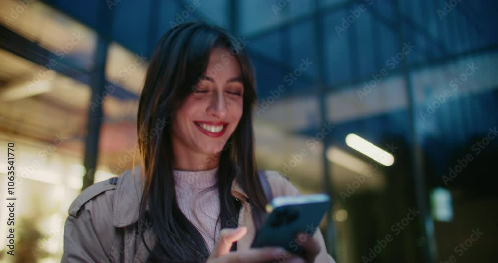 Beautiful Caucasian woman walking away from business center with tall glass windows. Smiling with joy after reading funny notification or message from friend. Leaving work late in evening.