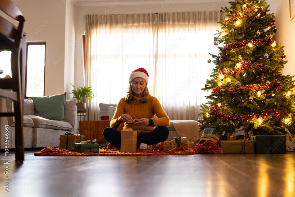 senior woman wrapping gifts by Christmas tree at home, wearing Santa hat, smiling senior woman wrapping gifts by Christmas tree at home, wearing Santa hat, smiling