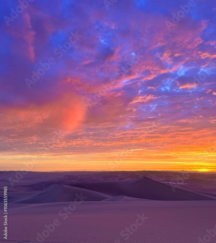 A vibrant sunset over desert dunes with a sky in shades of pink, purple, and orange. The warm colors cast a serene glow over the vast, tranquil landscape, creating a peaceful scene.