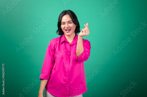 An Asian woman in a pink shirt stands against a green background, smiling warmly as she makes a Korean finger heart gesture with one hand, symbolizing affection, positivity, and friendliness