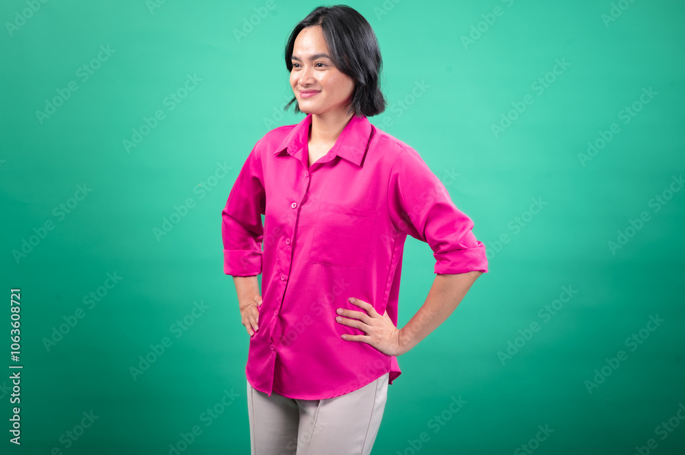 An Asian woman in a bright pink button-up shirt stands confidently with her hands on her hips against a solid green background, projecting a friendly and assertive expression An Asian woman in a bright pink button-up shirt stands confidently with her hands on her hips against a solid green background, projecting a friendly and assertive expression