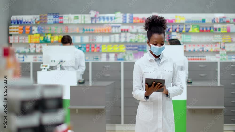 African American female pharmacist holding tablet at foreground. Woman druggist working with gadget at workplace. Woman in white robe and mask near showcase with medicine. Ethnic diversity. African American female pharmacist holding tablet at foreground. Woman druggist working with gadget at workplace. Woman in white robe and mask near showcase with medicine. Ethnic diversity.