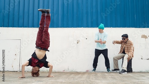 Young street dancer dancing in hip hop style with multicultural friends cheering behind at wall. Attractive handsome man stretch arms at street with blue background. Outdoor sport 2024. Hiphop. photo