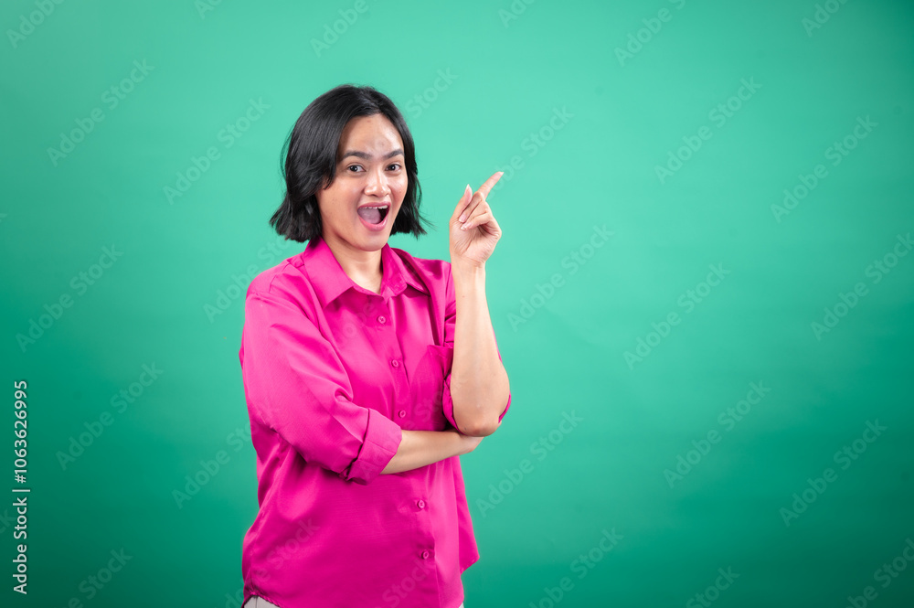 An Asian woman in a vibrant pink shirt stands against a green background, looking cheerful and pointing upwards with one hand, as if indicating a bright idea or something exciting An Asian woman in a vibrant pink shirt stands against a green background, looking cheerful and pointing upwards with one hand, as if indicating a bright idea or something exciting