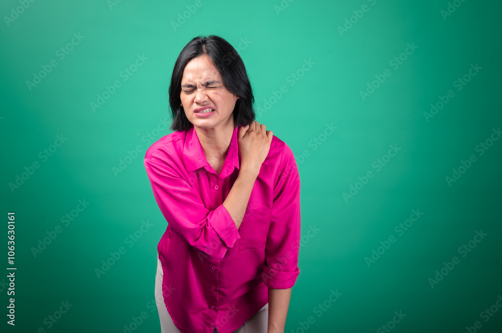Fototapeta premium An Asian woman wearing a bright pink shirt against a green background is holding her shoulder with one hand, expressing pain or discomfort, with her face showing a pained expression
