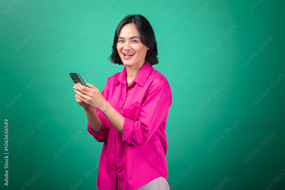 An Asian woman in a bright pink shirt smiles while looking down at her smartphone, holding it with both hands as if interacting with a game or reading something exciting. Her expression is cheerful An Asian woman in a bright pink shirt smiles while looking down at her smartphone, holding it with both hands as if interacting with a game or reading something exciting. Her expression is cheerful