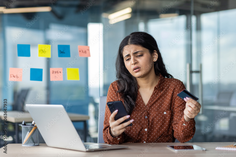 Young Hispanic businesswoman looks concerned while using phone and credit card in office. Technology, finance, stress concepts portrayed as professional engages with smartphone, laptop, tasks. Young Hispanic businesswoman looks concerned while using phone and credit card in office. Technology, finance, stress concepts portrayed as professional engages with smartphone, laptop, tasks.