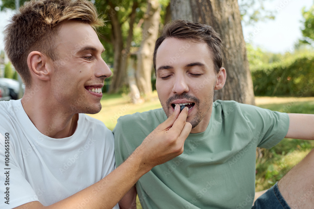 Fototapeta premium Two smiling men enjoy a playful moment while feeding each other outdoors in a beautiful setting.