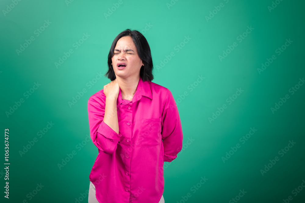 Fototapeta premium An Asian woman wearing a bright pink shirt against a green background is holding her shoulder with one hand, expressing pain or discomfort, with her face showing a pained expression