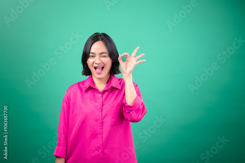 An Asian woman in a bright pink shirt poses against a green background, smiling and winking while making an "OK" gesture with her hand. Her expression is cheerful and playful