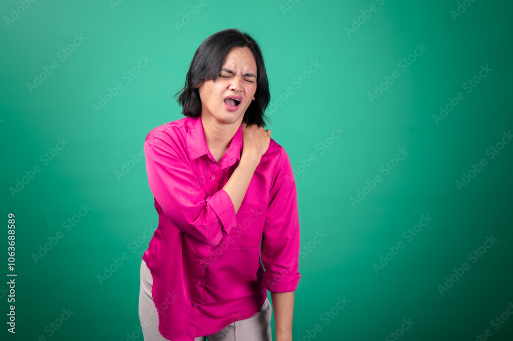 Fototapeta premium An Asian woman wearing a bright pink shirt against a green background is holding her shoulder with one hand, expressing pain or discomfort, with her face showing a pained expression