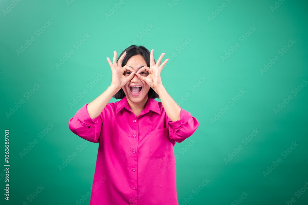 An Asian woman in a bright pink shirt poses against a green background, smiling and winking while making an An Asian woman in a bright pink shirt poses against a green background, smiling and winking while making an