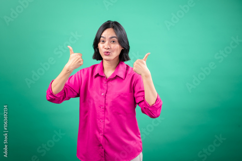 An Asian woman in a vibrant pink shirt stands against a green background, smiling and giving a thumbs-up gesture. Her cheerful expression and confident pose convey positivity and approval.