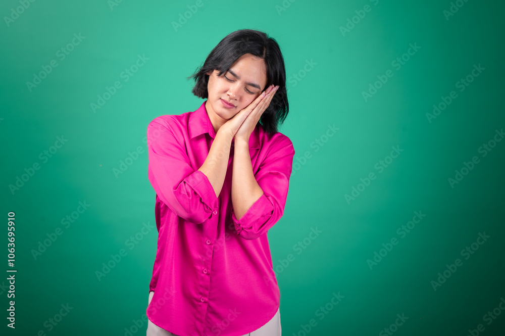 An Asian woman in a pink shirt stands against a green background, holding her hands together near her cheek in a sleeping gesture. Her eyes are closed, expressing calmness and a peaceful mood. An Asian woman in a pink shirt stands against a green background, holding her hands together near her cheek in a sleeping gesture. Her eyes are closed, expressing calmness and a peaceful mood.