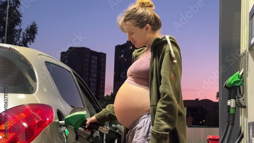 Young pregnant woman fills up her car at a gas station in the evening