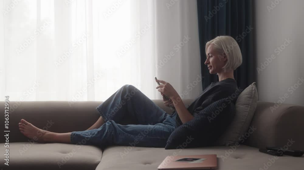 Attractive young blonde woman relaxing on her couch using her phone and smiling. Laptop and TV remote nearby. Natural light. Cozy home. Attractive young blonde woman relaxing on her couch using her phone and smiling. Laptop and TV remote nearby. Natural light. Cozy home.
