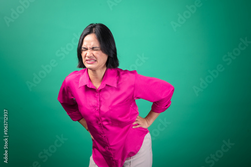 An Asian woman in a bright pink shirt against a green background expresses pain, with her hand on her back as if experiencing backache or discomfort. Her face shows an intense expression of distress.