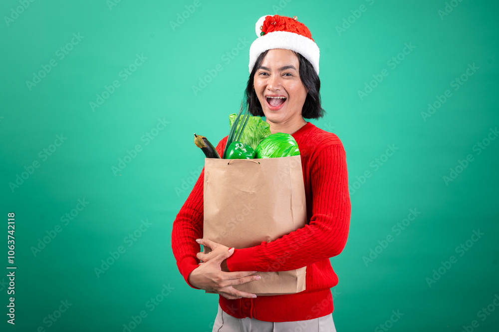 Asian woman wearing a Santa hat and red sweater smiles joyfully while holding a brown paper bag filled with fresh green vegetables against a green background, embracing the holiday season Asian woman wearing a Santa hat and red sweater smiles joyfully while holding a brown paper bag filled with fresh green vegetables against a green background, embracing the holiday season