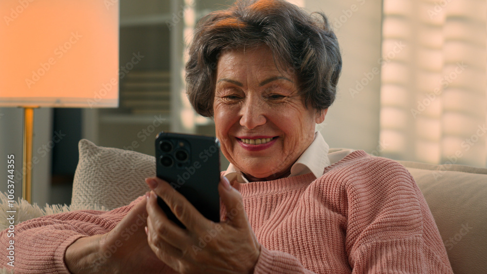 Fototapeta premium Happy smiling carefree laughing Caucasian old woman mature lady retired female senior granny grandmother chatting with smartphone watching funny video reading good news browsing mobile phone at home