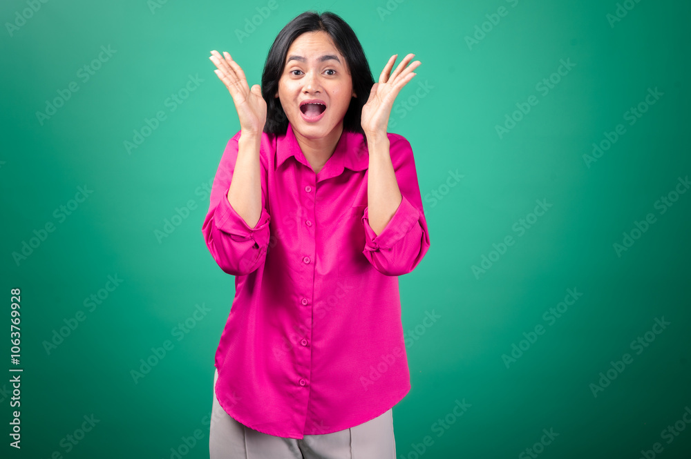 Fototapeta premium An Asian woman in a bright pink shirt stands against a green background, expressing surprise with an open-mouthed smile, her hand raised to her face in a playful, animated gesture