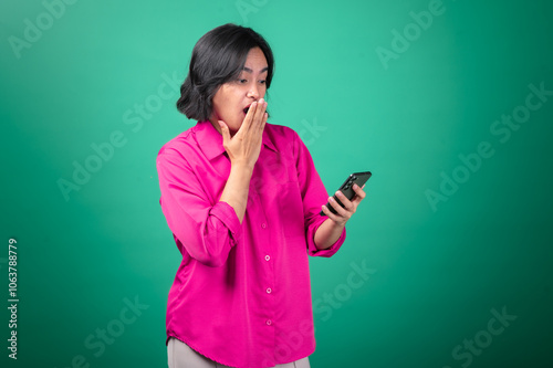 An Asian woman in a bright pink shirt holds a smartphone in one hand, covering her mouth in surprise with the other. Her expression shows shock and curiosity.