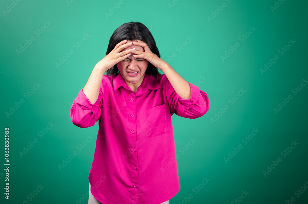 An Asian woman in a pink shirt stands against a green background, with her hands on her head and her mouth open, expressing frustration or stress. Her face shows an intense, emotional reaction An Asian woman in a pink shirt stands against a green background, with her hands on her head and her mouth open, expressing frustration or stress. Her face shows an intense, emotional reaction