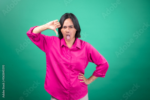 An Asian woman in a bright pink shirt poses against a green background, making a playful face by sticking out her tongue and giving a thumbs-down gesture. Her expression is humorous