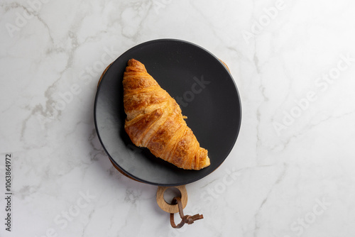 A freshly baked golden croissant rests on a sleek black plate, set against a faintly textured marble background, highlighting its perfect flakiness and appetizing form. photo