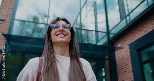 Charming Caucasian girl with glasses and straight hair walking past modern building with brick walls and tall windows. Smiling while looking straight ahead. Going to planned event for evening.