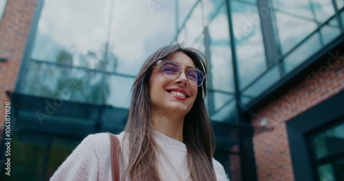 Positive Caucasian woman confidently walking away from office building. Leaving work early. Smiling while looking around. Happy about upcoming weekend. Wearing round glasses and handbag. photo