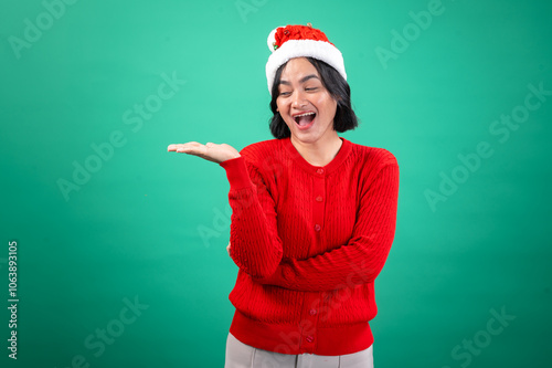 An Asian woman wearing a red sweater and Santa hat, smiling widely with one hand raised in a welcoming gesture, standing against a green background. She exudes festive joy and holiday cheer