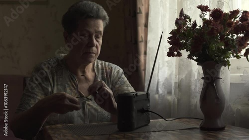 Elderly woman tuning and listening to portable radio while sitting by window.