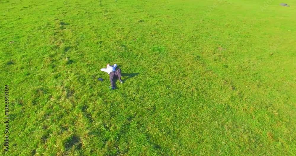Aerial view point. Low orbital flight around man lied on green grass with notebook (pad) at yellow rural field. Casual weared. Sun rays. Radial movement. Aerial view point. Low orbital flight around man lied on green grass with notebook (pad) at yellow rural field. Casual weared. Sun rays. Radial movement.