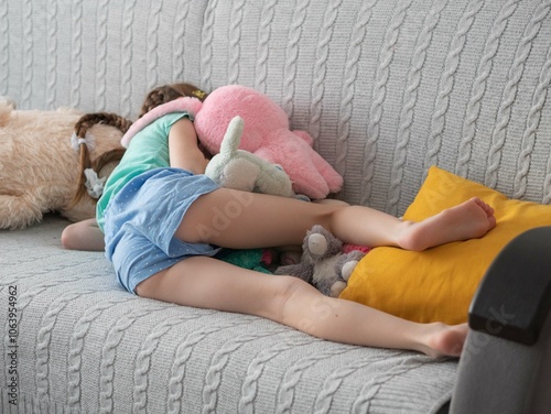 A little upset girl lies on the couch in a pile of soft toys, turned away, hides a tear-stained face, bare feet photo