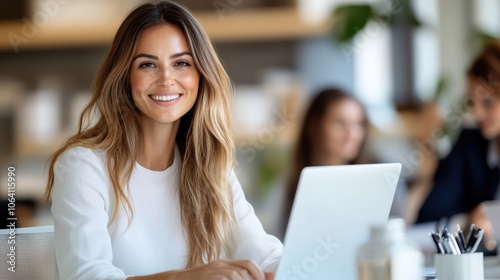 A woman with long hair is smiling while working on her laptop in an office full of natural light, indicating productivity, positivity, and a modern workspace.